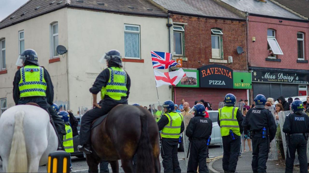 Mounted police and officers wearing riot gear line a street while protesters march through the high street waving Union Flags.