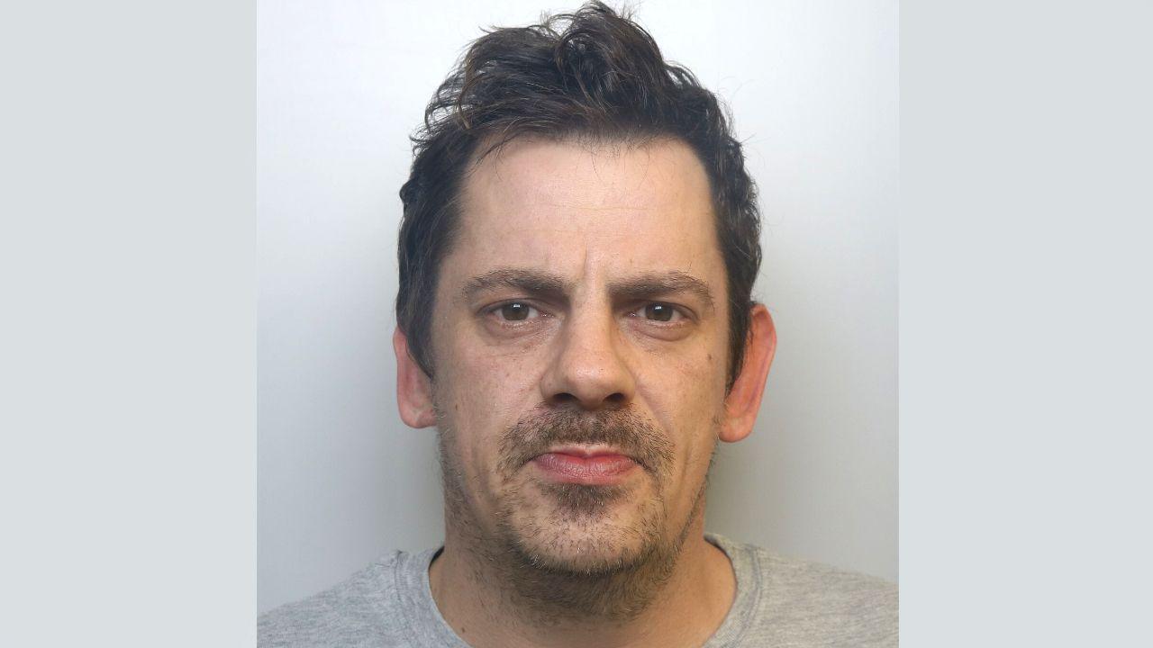 A police mugshot of Mark Roberts. He is wearing a grey t-shirt and standing against a grey background. He has dark hair which is styled upwards at the front, dark eyes and a patchy goatee style beard. He is looking at the camera with a blank expression. 