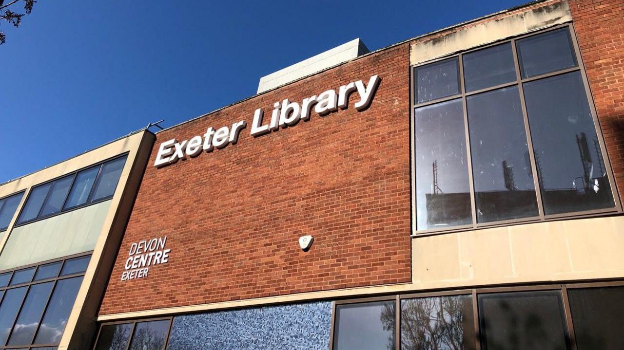 The sign for Exeter Library on the front of the building in white against red brick and with panelled windows on either side.