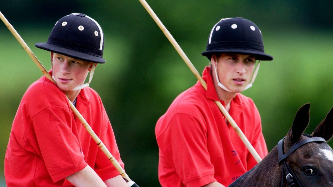 Prince William and Prince Harry on horseback at a polo match in 2002. They are both wearing full polo kits, red shirts and mallet over their right shoulder.