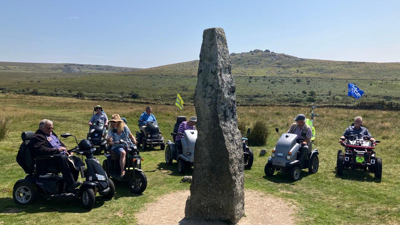 Eight people in mobility scooters in a semicircle around a large triangular rock. The grass is green. There is a hill in the background. The sky is a light blue. 