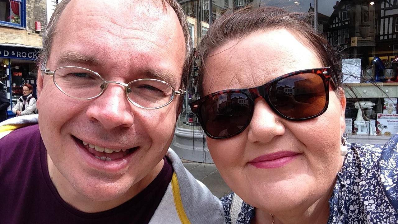 A selfie of Joy Beard and her husband James. She is wearing dark sunglasses and a blue shirt. James is wearing normal glasses and has a grey hoodie on over a maroon top