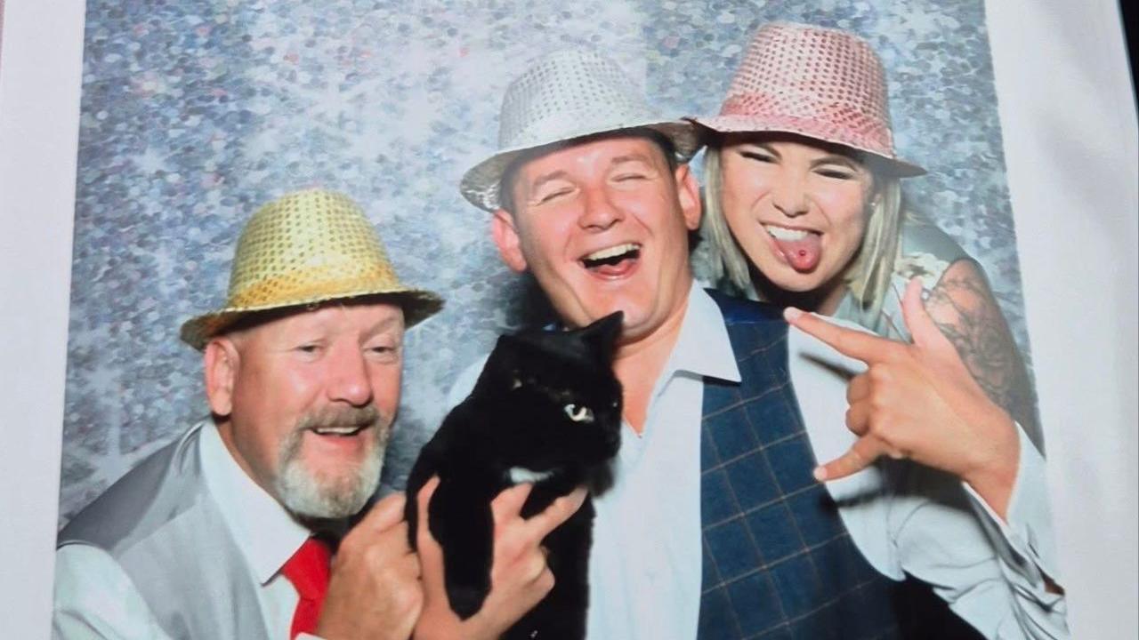 A picture of two men holding Polo the cat at a wedding. There is a woman on the right poking her tongue out. The man in the middle is smiling as well as the man on the left. Polo is looking to the right.