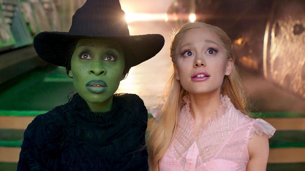 Cynthia Erivo and Ariana Grande stare into the camera dressed as their Wicked characters Elphaba and Glinda, in a scene from the film Wicked. Elphaba wears a black outfit and black witch's hat, while Glinda wears a pink dress.