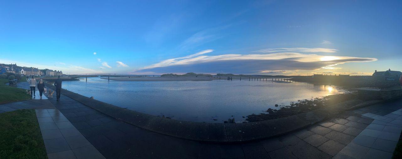 A panoramic photo of a harbour with a blue sky above and three people walking at the left-hand side