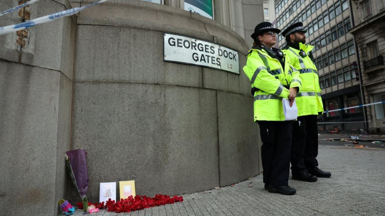 Police officers stand next to flowers and cards placed on the ground near the site of an incident where a car plowed into a crowd of Liverpool fans during a parade celebrating their side's Premier League soccer title, in central Liverpool.