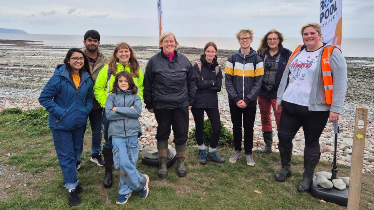 A team photo of people taking part in the Klive beach rock pool challenge. They are standing by a rocky beach, with the grey sea behind them.