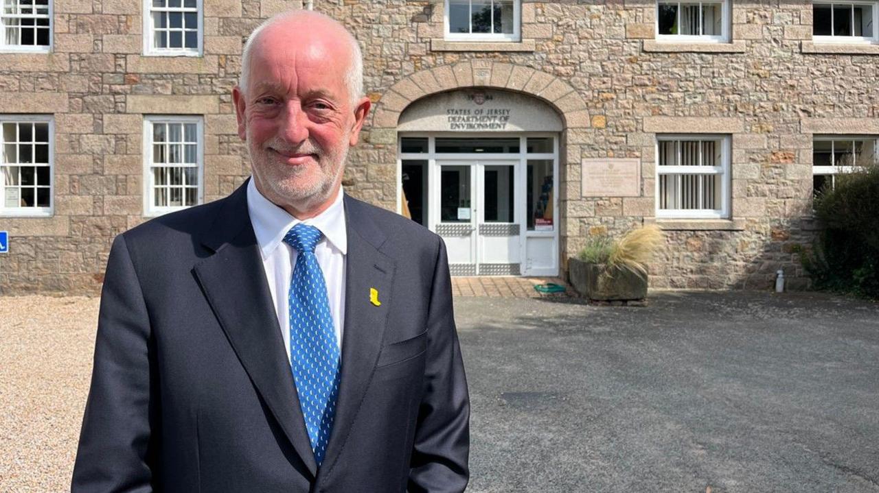 Deputy Steve Luce, in a dark suit, blue and white patterned tie and a white shirt, standing outside the main entrance to Jersey's Environment Department at Howard Davis Farm.