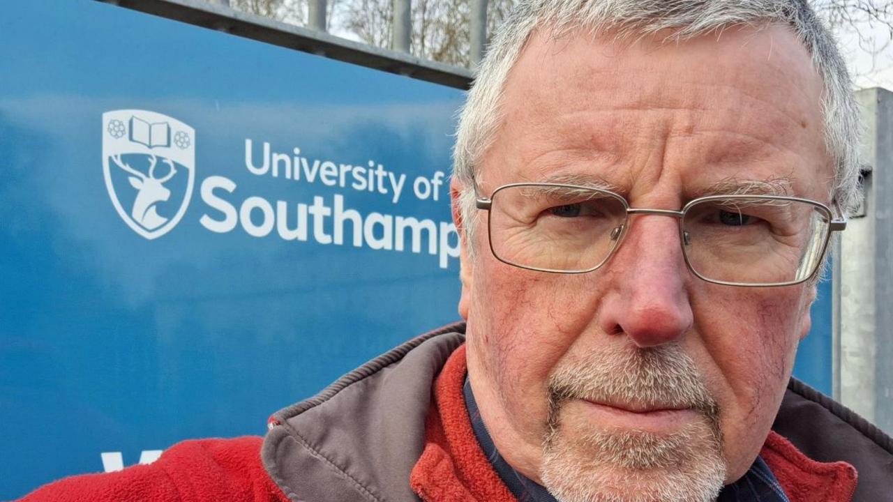 A photograph of Stephen Hanvey in the foreground, with the University of Southampton written in white text on a blue sign in the background, next to an image of a shield with stag inside of it. 