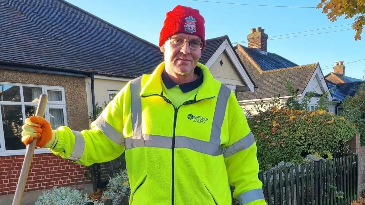 Wayne Broadhurst is pictured wearing high visibility uniform with gloves and a hat while holding a broom stick. Houses are visible in the background.