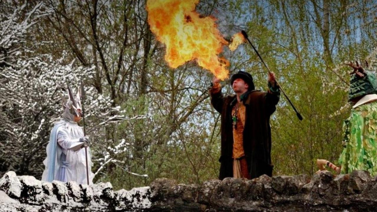 A fire-breather wearing a hat and a dark coat is standing near two people, who are dressed up, either side of him. Trees are in the background.