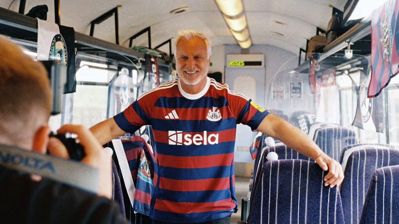 David Ginola, a tall man with white hair and a white beard, stands in a train carriage smiling wearing the blue and red Newcastle United away kit. In the foreground, George Dixon is taking his picture on a film camera.