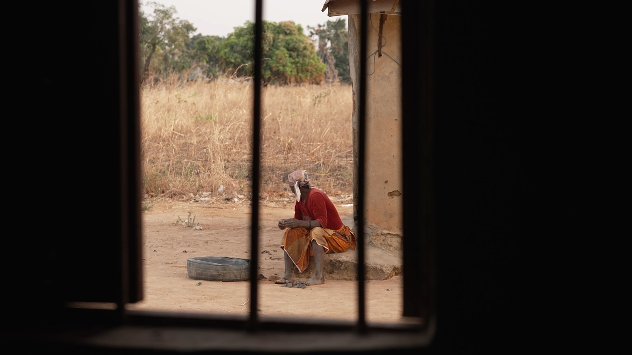 Sarah Peter sitting on a concrete slab. She is seen in the distance and photographed through the bars on a window. She is turning away from the camera.
