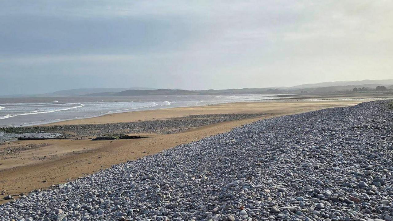 A screenshot from Google street view showing Dunster Beach at low tide. It is a vast shingle beach with with a large bay and headlands opposite.