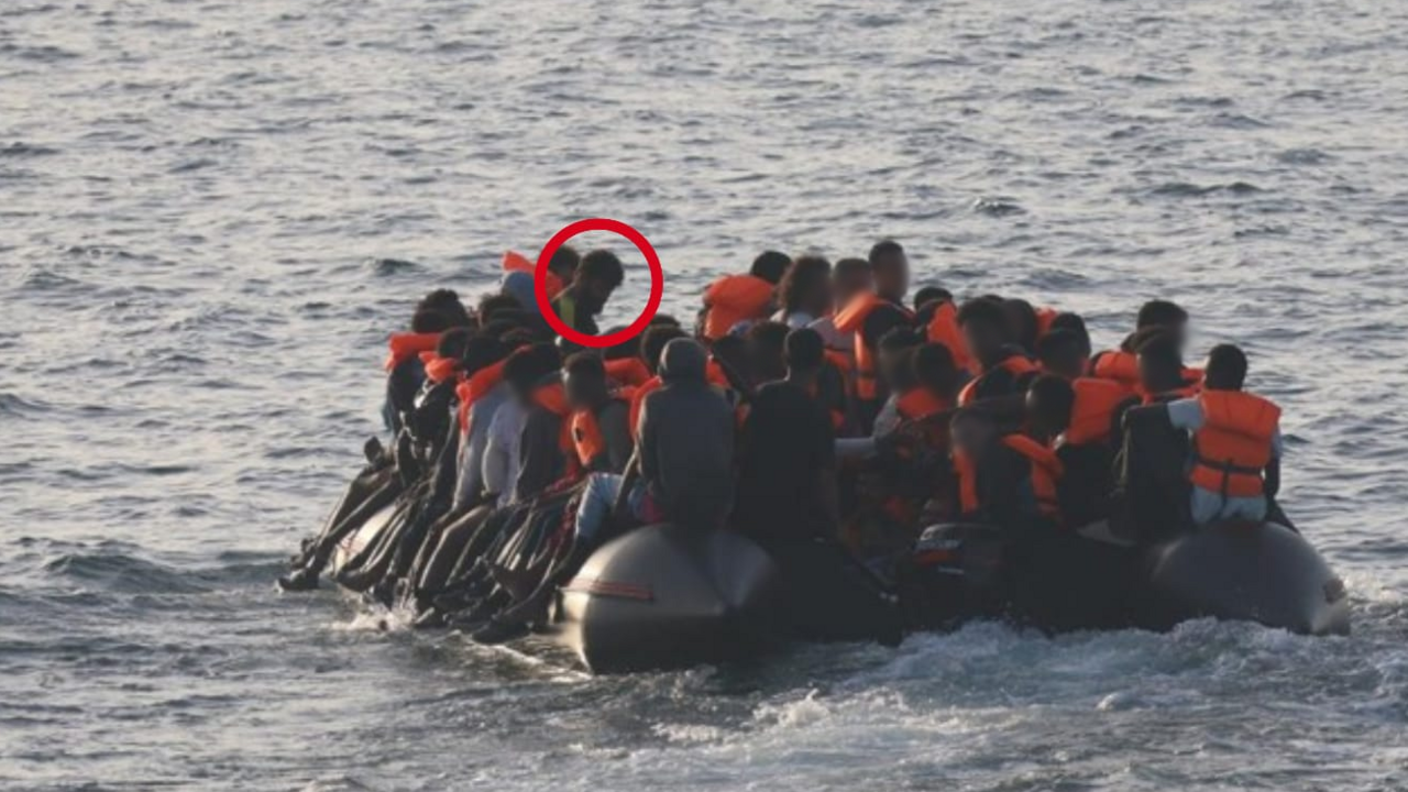 An image of an overcrowded dinghy at sea. Dozens of figures can be seen, with some wearing orange life vests. There are waves in the water, which is grey, and the people have their backs to the camera.