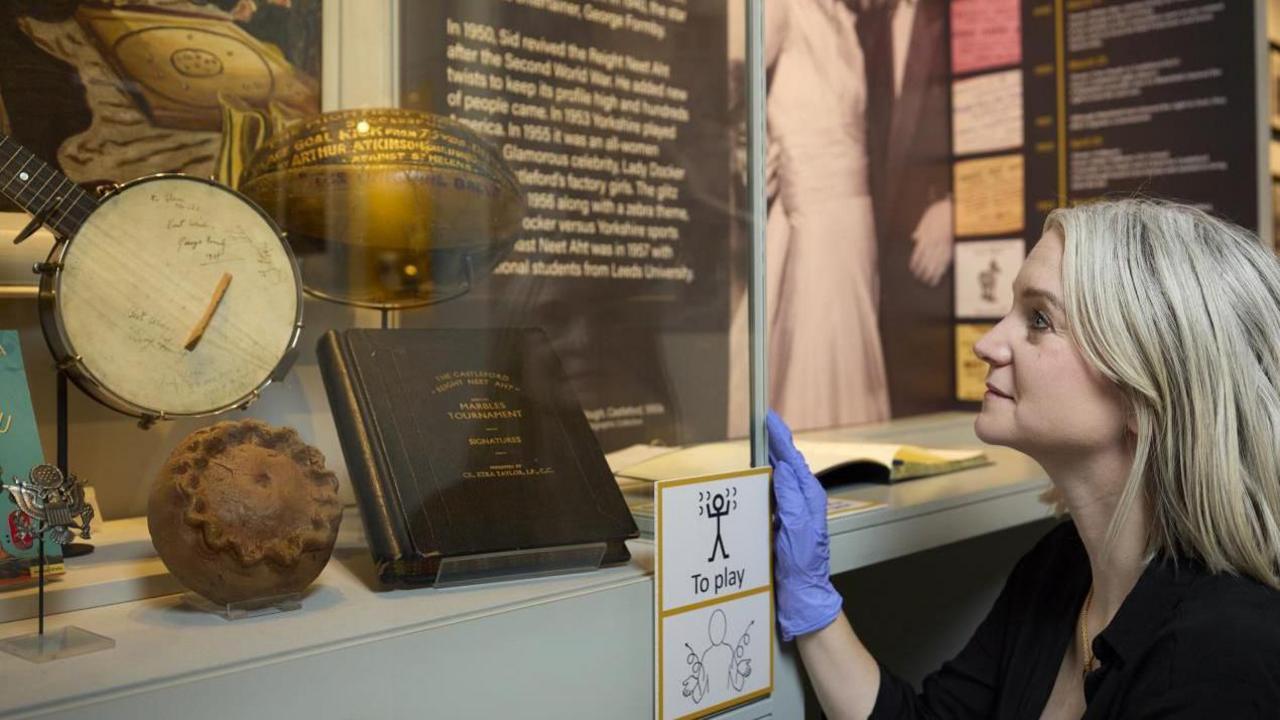 A blonde woman looks at objects at Castleford Museum wearing blue gloves