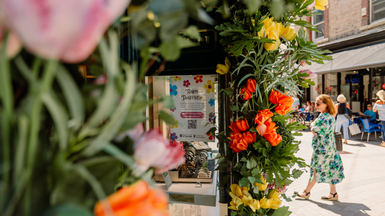A floral display on the left with a sign at the centre. There is a woman walking towards the flower display, and people sat down at a cafe in the distance.