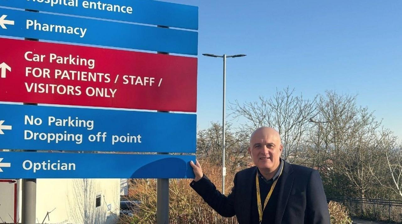 Councillor Ed O'Driscoll wearing a dark jacket, is facing the camera, touching a large sign outside Ross-on-Wye Community Hospital. The red and blue sign has white arrows pointing to various places at the site.