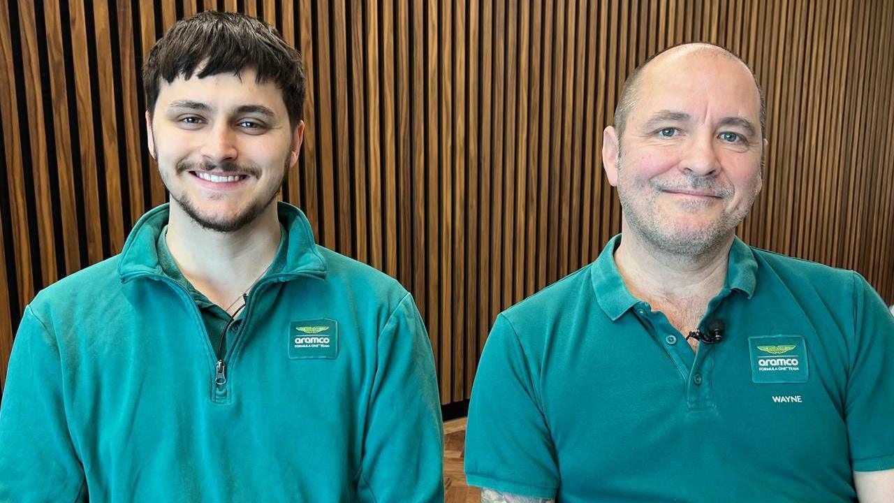 Wayne Meakin and his son Cole Fell-Meakin, sitting and smiling and wearing green shirts. They are in an office environment with a wooden background.