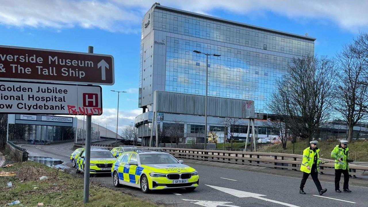 Two police officers in fluorescent yellow jackets walk towards the scene of a serious road accident. Four police cars are in the background on the deserted road. A large office block can be seen in the right of the image while road signs are visible in the foreground. 