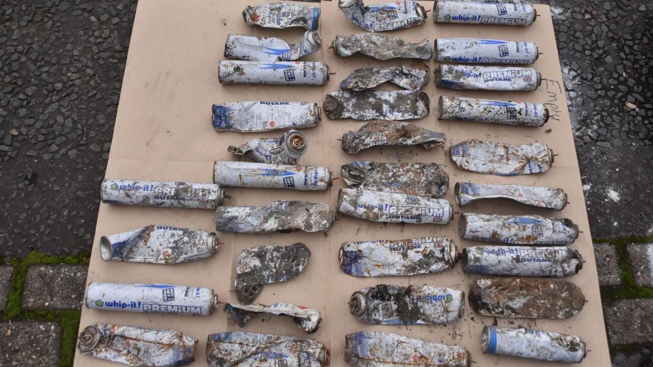 34 butane canisters laid out on a board of wood. They are in varying conditions, some just reduced to ragged metal, others still showing the white and blue and distinctive cylindrical shape