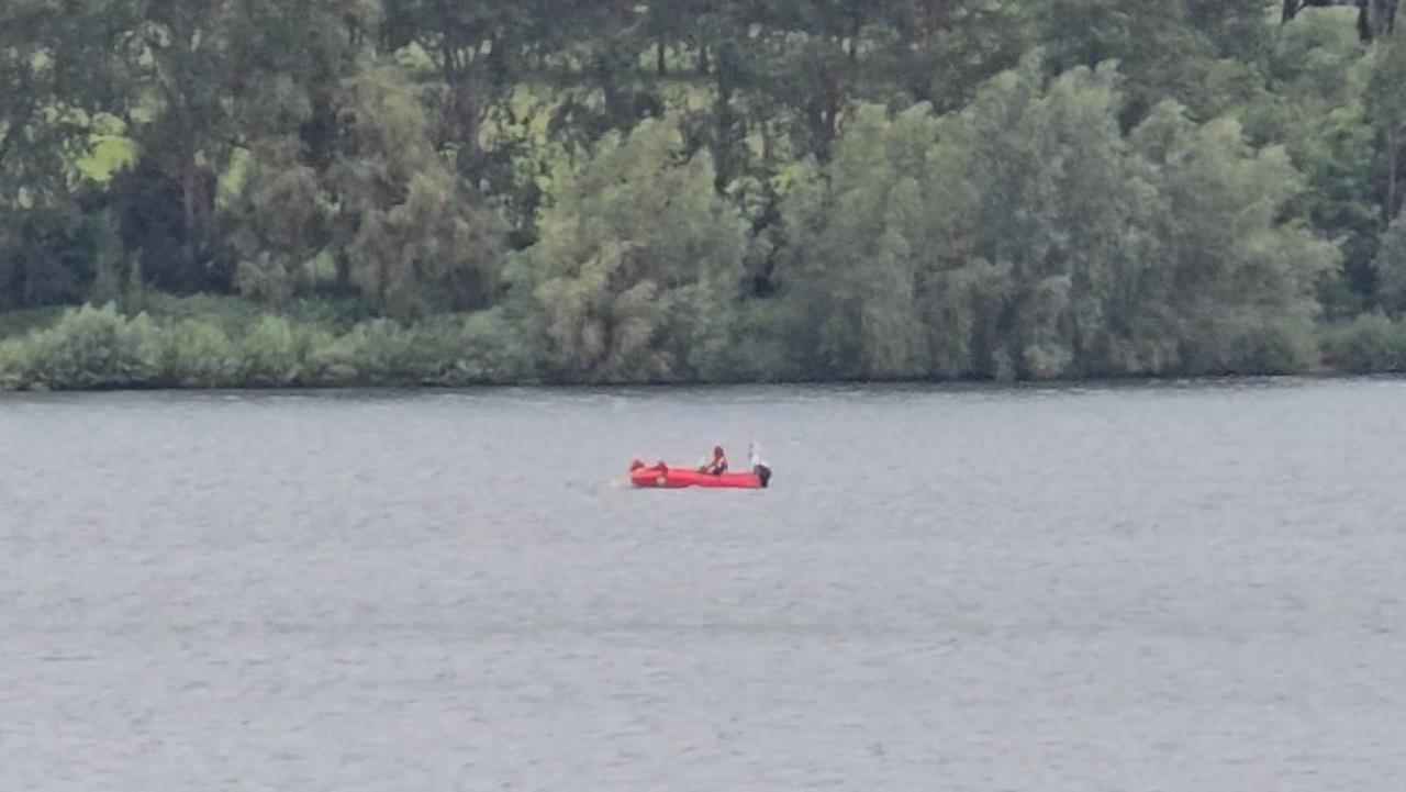 A red rescue boat with a person in it on a reservoir. Trees are seen behind the water.