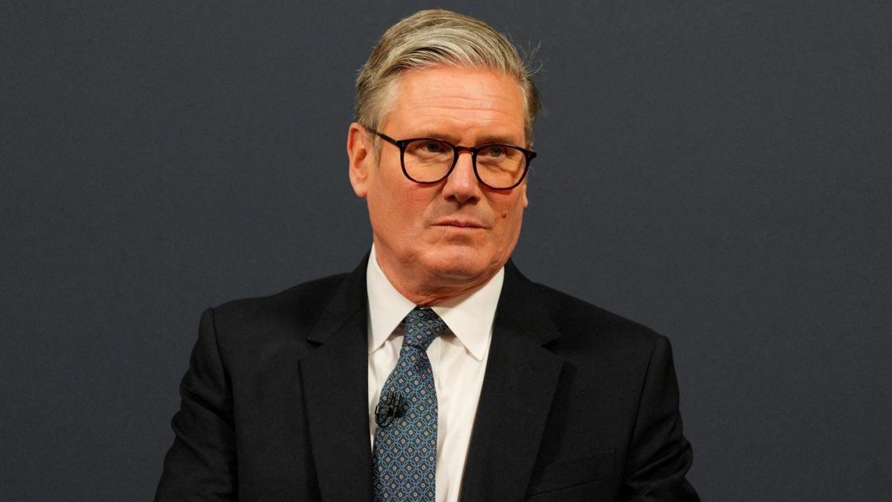 Sir Keir Starmer has grey hair which is slicked back to one side of his head with shorter sides and is wearing oval glasses, a black suit jacket, white shirt and blue tie with yellow triangular print on it. He has a microphone attached to his tie and is looking to the side with a serious expression. The background is dark grey.