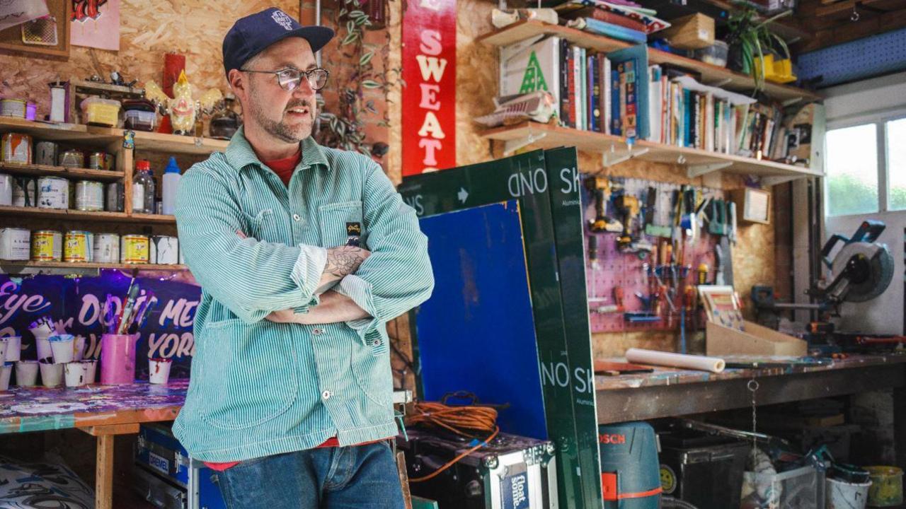 A man wearing a blue cap and a green and white striped shirt over a red t-shirt with jeans, stands in a workshop surrounded by paint and painted panels. There are bookshelves in the background and he is wearing glasses and has his arms folded. 