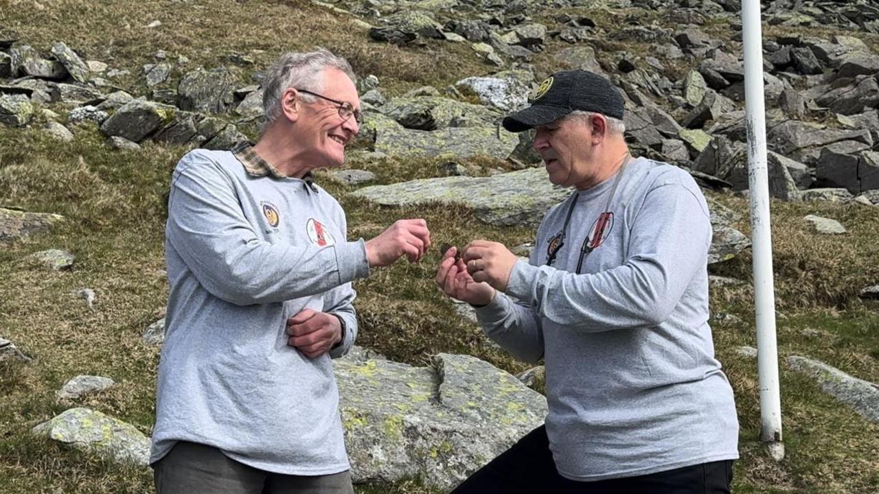 Ivor Ramsden (left) wearing a grey sweatshirt and glasses, passing the dog tag to Don Madar who is also wearing a grey sweatshirt with a black baseball cap. They're on the rocky hillside and there is a white flag pole to the right.