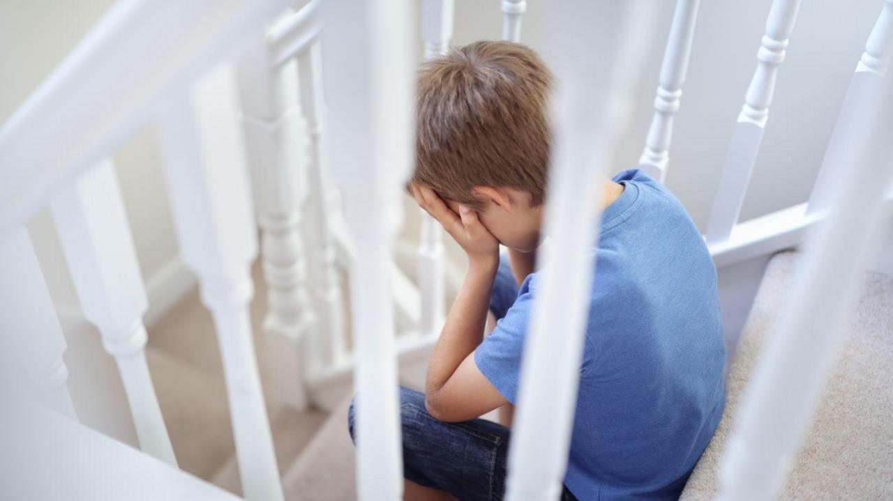 A boy with blonde hair and a blue tshirt sitting on stairs with white bannisters with his head in his hands