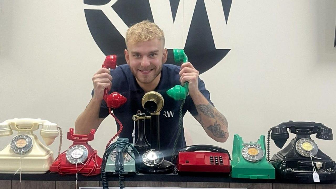 A man with blond hair is smiling at the camera and holding the receiver of a red phone in his right hand and of a green phone in his left hand. Seven phones are on the flat surface in front of him.