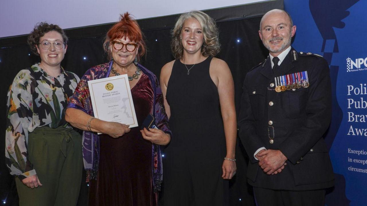 Four people - three women and a man - stand in a line smiling at the camera. Marion Balaam is pictured holding a certificate and wears a purple and red dress and shawl. Hannah and Gavin are wearing black and Emily wears green trousers and a green and white shirt.