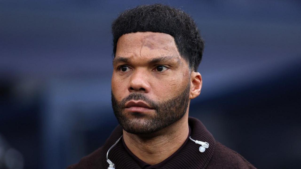 A man with dark hair and a beard is looking away from the camera. The blurred background is light blue.