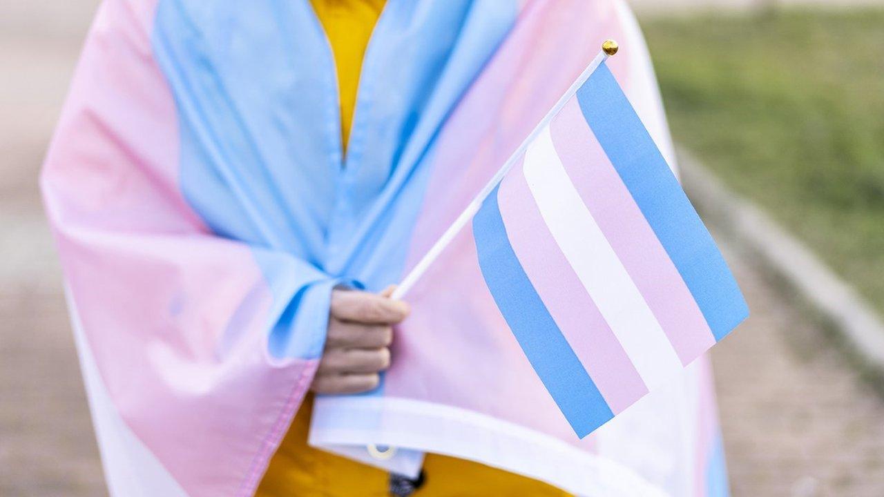 A young person wrapped in a pink, white and blue transgender pride flag