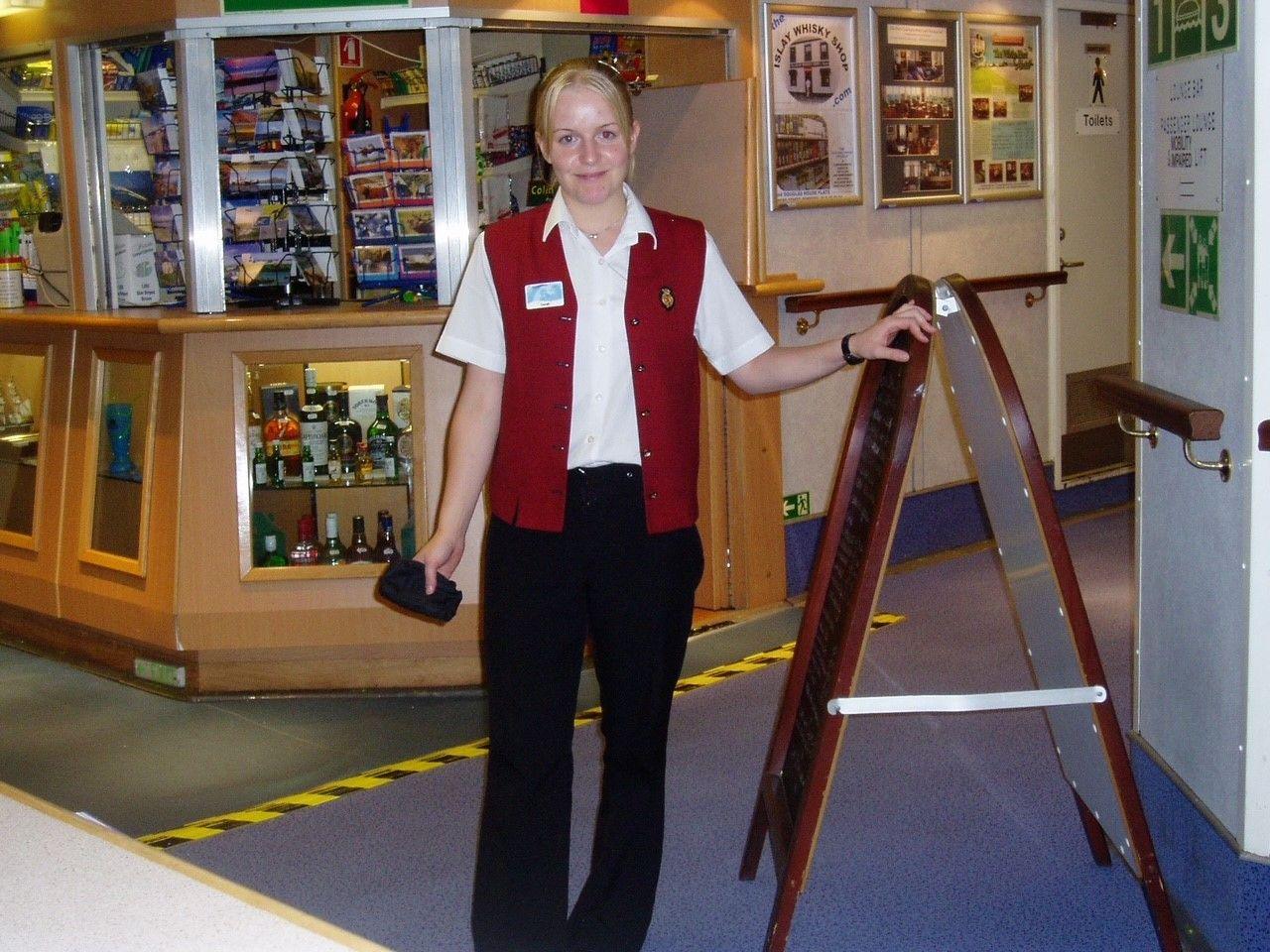 A blonde haired woman in black trousers, white shirt and red waistcoast stands in front of a gift shop on board a ship