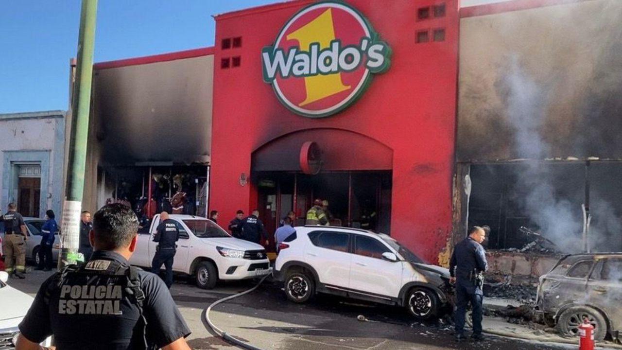  A building that says Waldo's on the front is covered with black burn marks, with smoke rising from some of the windows. Police and fire services are at the scene 