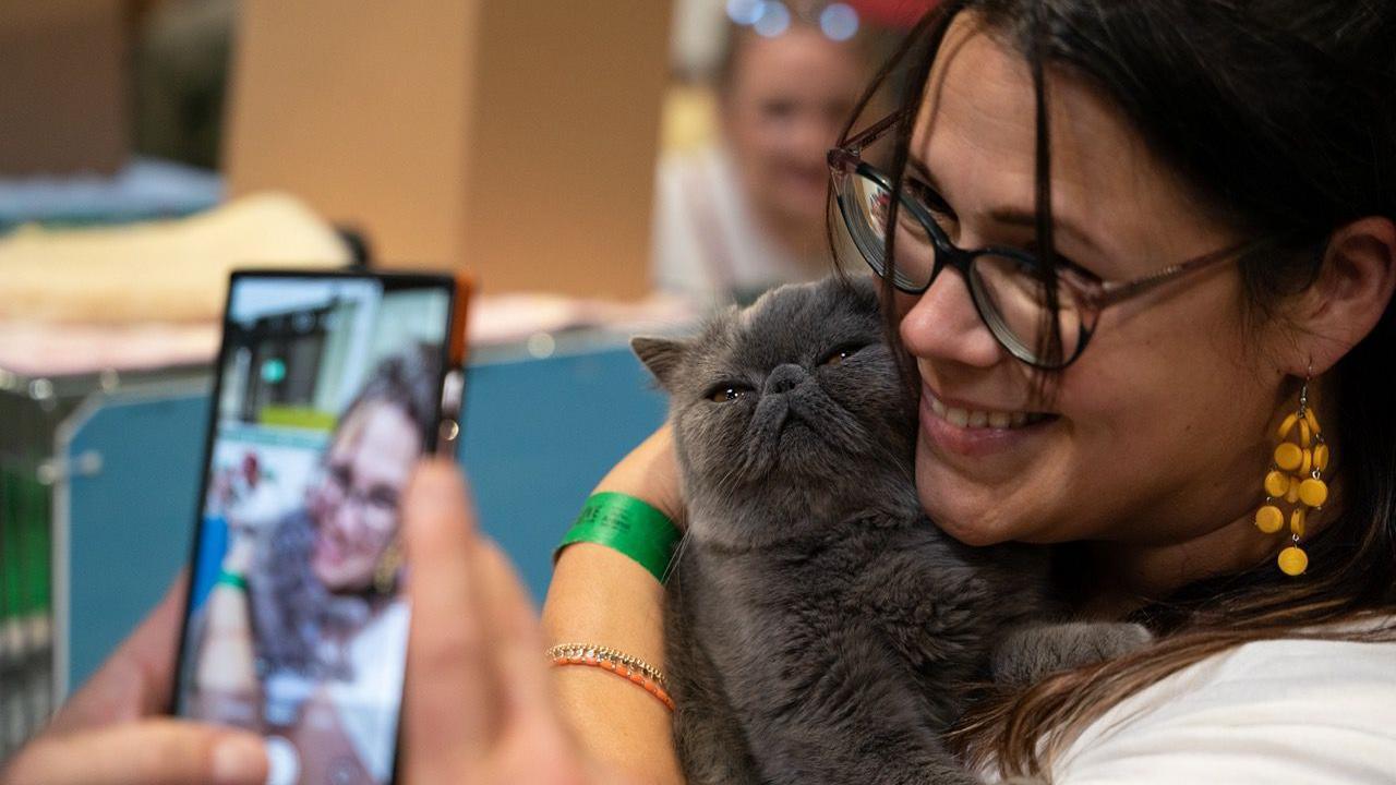 A woman with brown hair and glasses is hugging a cat while a phone takes a selfie of her smiling.