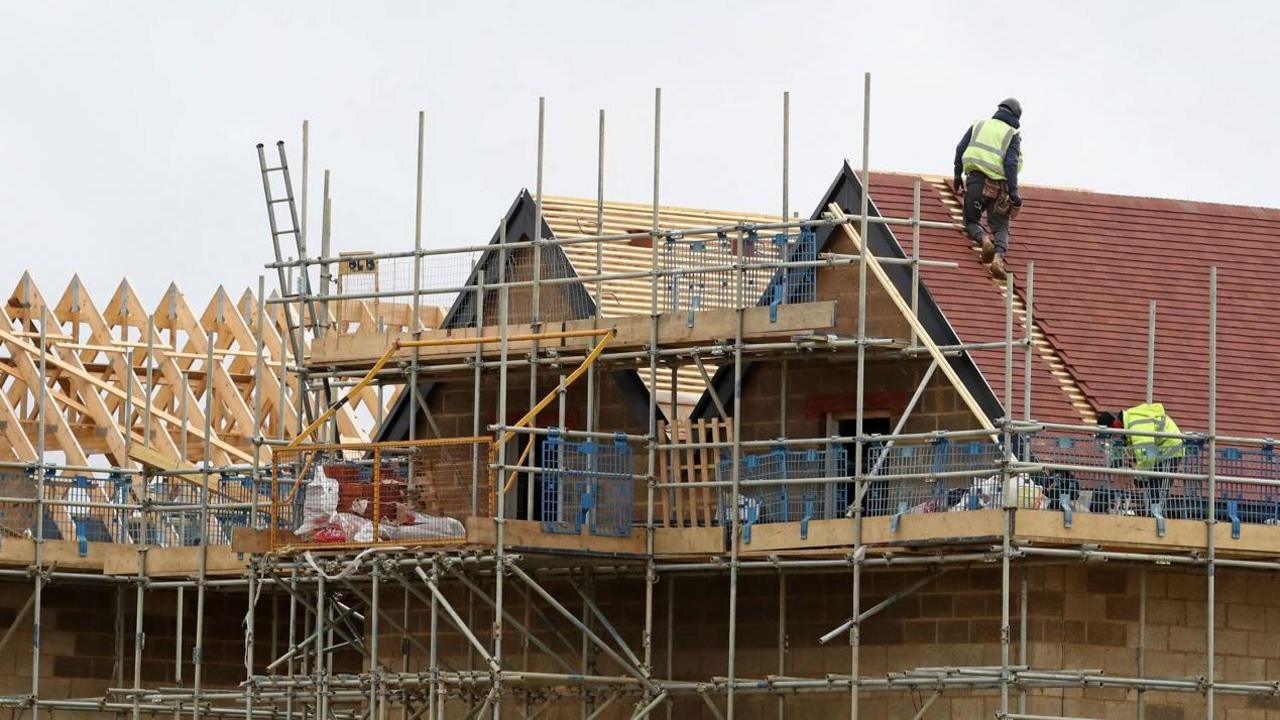 A stock image of two construction workers working on the roof of a half-built house with scaffolding around the outside