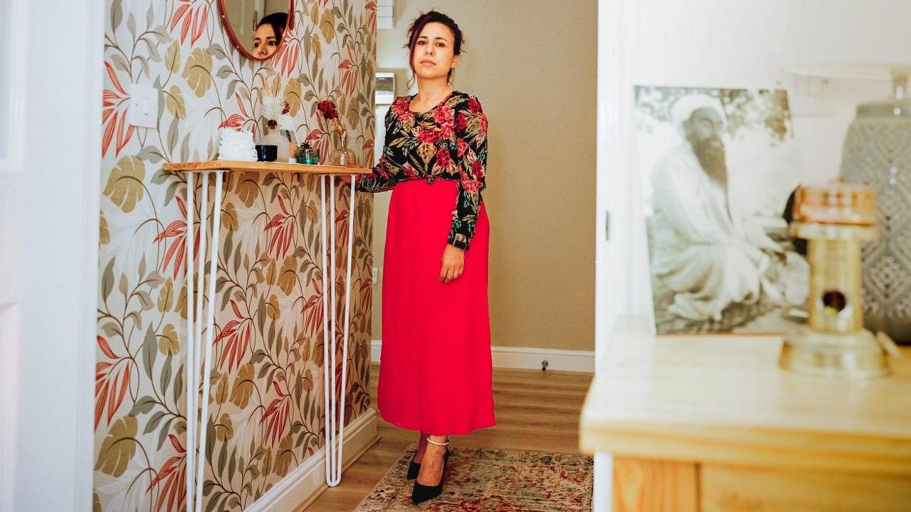 A lady wearing a red and black floral top and a long red skirt. She has her hand leaning on a shelf looking at the camera and in the foreground an old black and white photo of someone can be seen sat on a mantle piece.