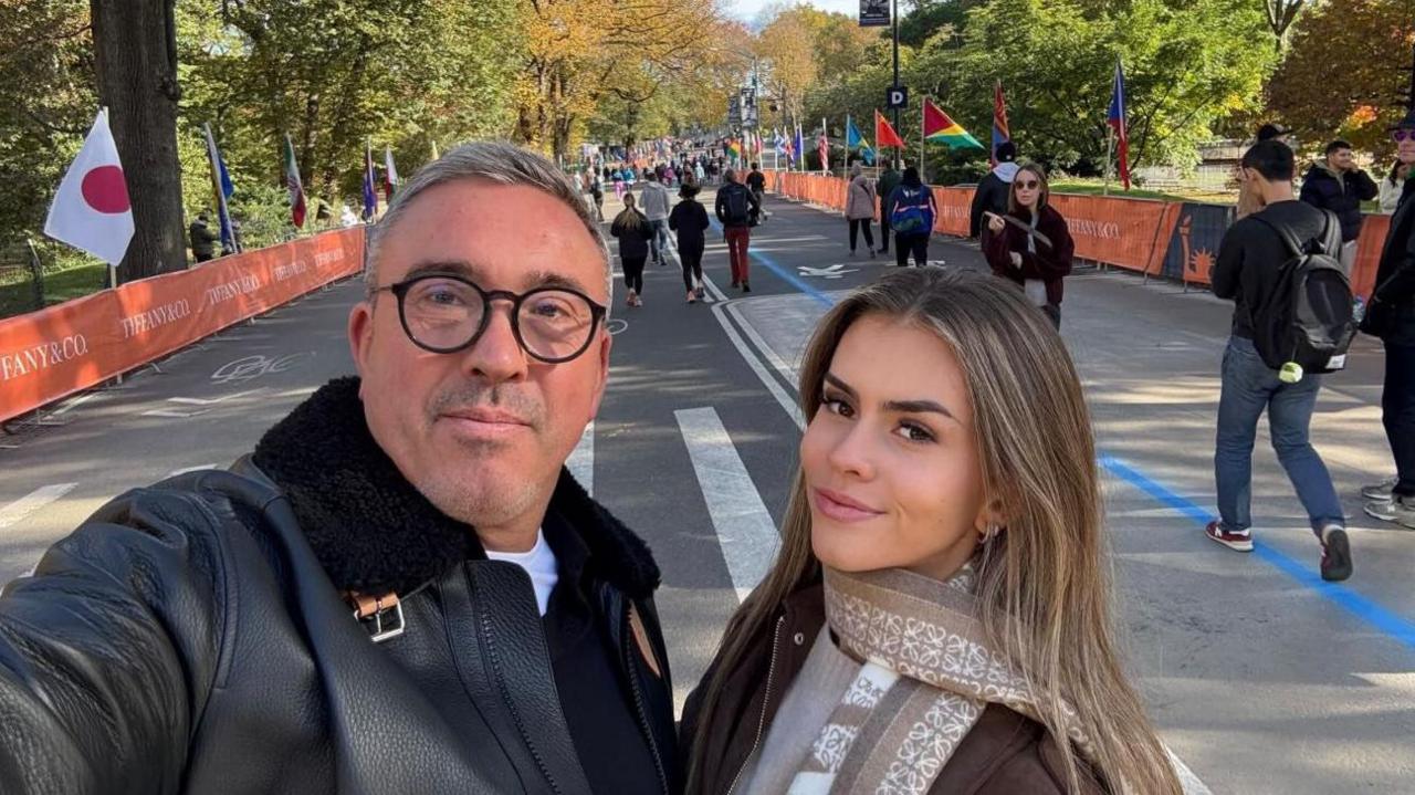 Lucy Scott and her dad John stand at the finish line of a race. Neither are competing. John is wearing a black jacket and has grey hair and black spectacles. Lucy is wearing a brown jacket with a beige scarf. They are both smiling at the camera. 