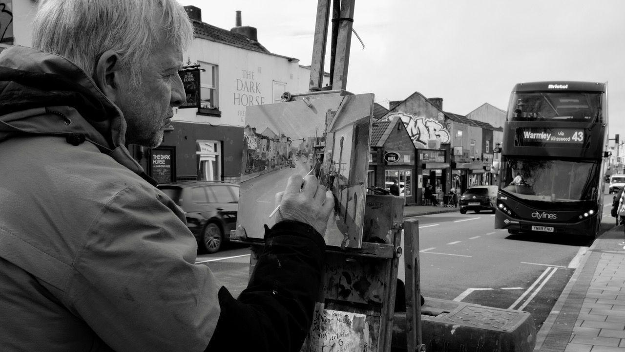 A black and white photo of artist Peter Brown painting a picture of a street. He is holding the paintbrush to the canvas and looking at his work. A bus heading for Warmley can be seen driving on the road ahead of him, and a pub and other businesses can be seen across the road. The photo is taken over Peter's shoulder, so only half his face can be seen.