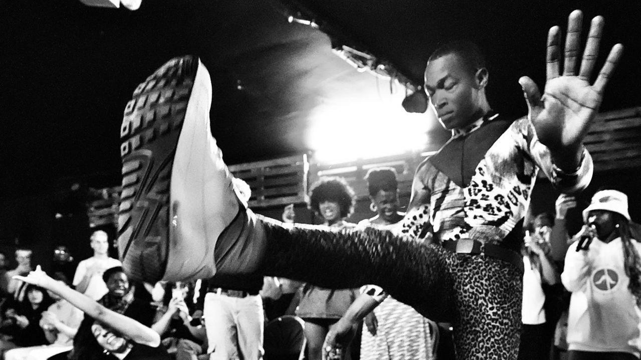 A man is dancing on stage. His foot is held up towards the camera. His eyes are closed. In the background a crowd is dancing and laughing.