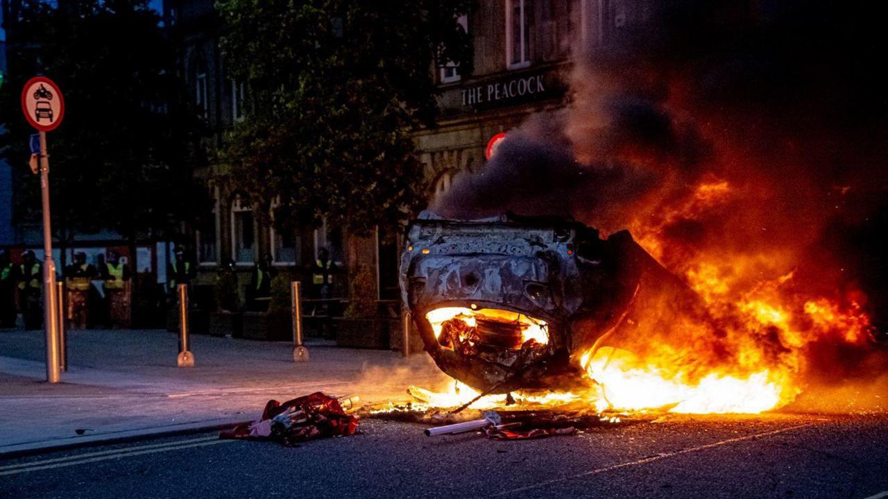 An overturned car engulfed in flames in the middle of a street in Sunderland. The Peacock pub is in the background while riot police are stood further back and to the left.