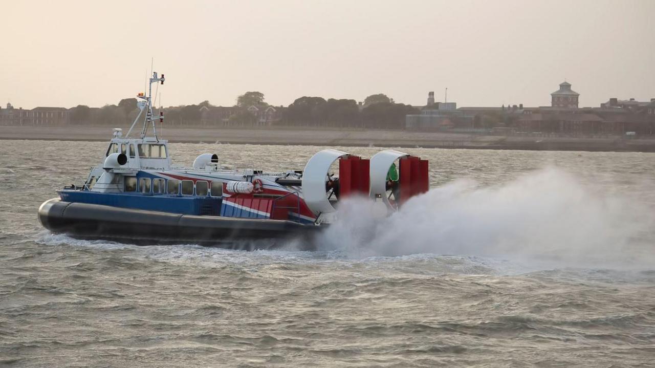 A side photo of a blue, red and white hovercraft in action off the English coast