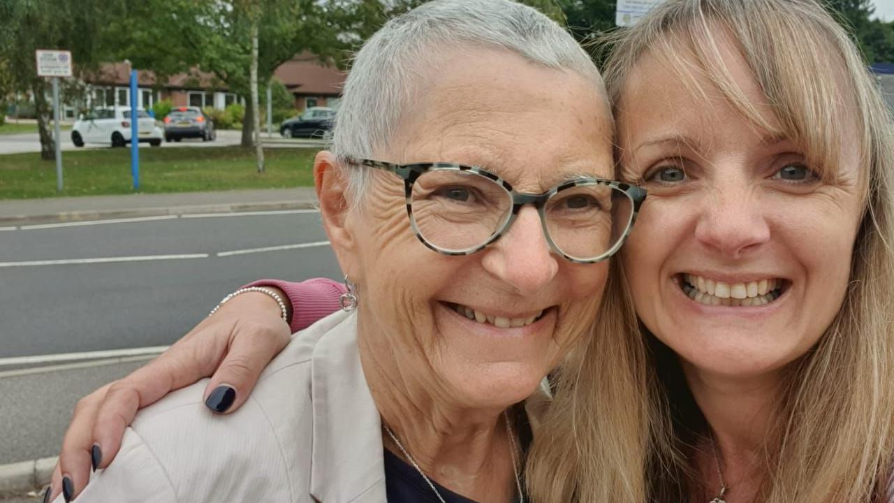 Gail Hardy (right) has her arm around her now late mum Sandra. Sandra left has short grey hair and wears glasses with a beige jacket. Gail has long blond hair. They are standing in the street with a road and car park behind them. Both are smiling. 