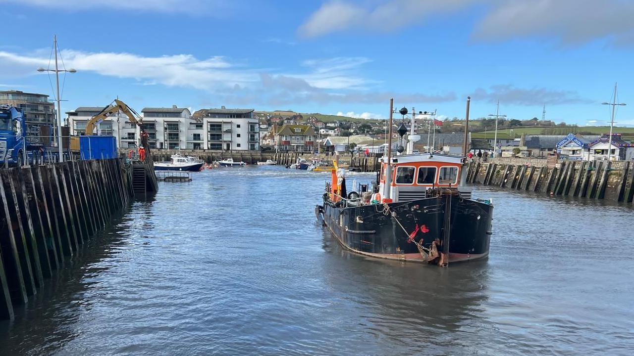 Front view of dredging vessel Doonhammer in Bridport harbour. The harbour walls rise up on both sides of the picture. In the background are three white modern blocks of flats with grey metal balconies.