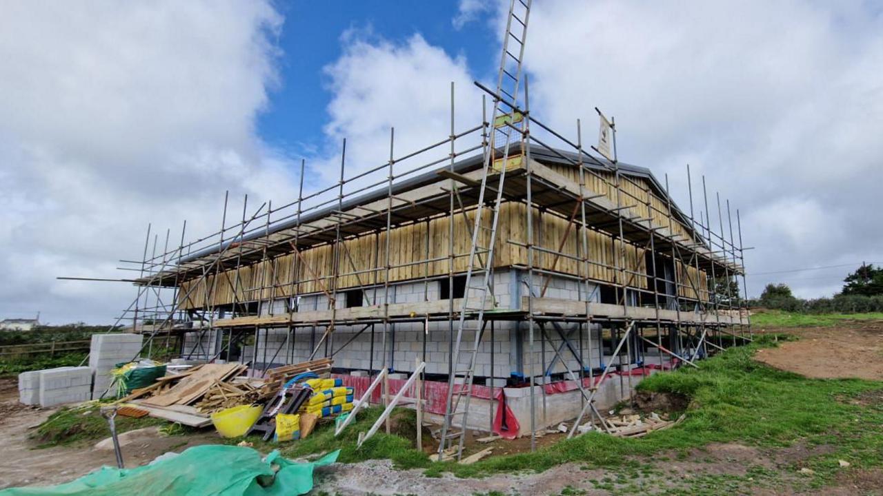 A building being constructed. It is surrounded with scaffolding. Bare breeze blocks are visible and the top half of the building is made of wood.