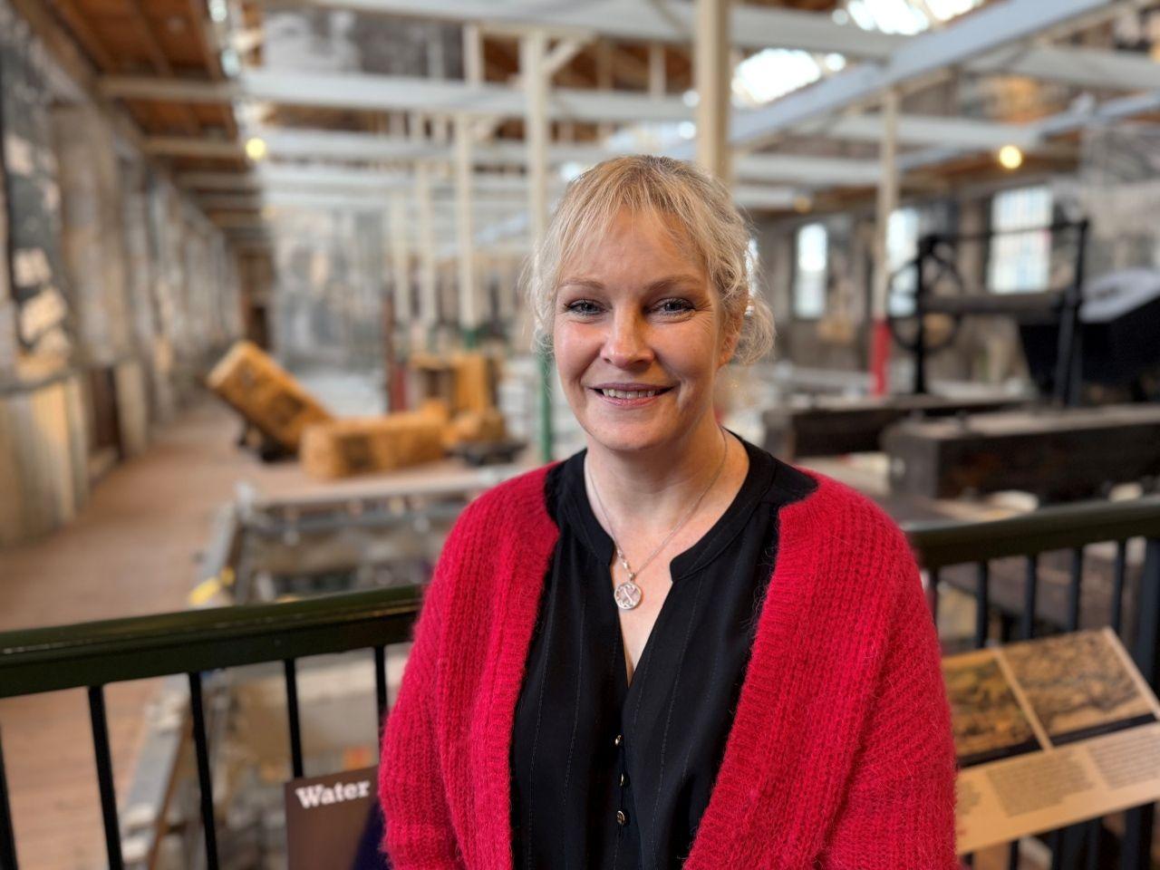 A woman with blonde hair, wearing a red cardigan and black top. She is standing in a museum with exhibits visible behind her