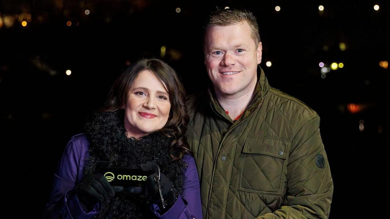 Vicky and her husband Dale standing outside at night with blurry lights in the background. She wears a purple raincoat and large black scarf, and holds a small black pouch with the word Omaze on it. Dale wears a green quilted jacket and has his arm round Vicky. He has cropped light brown hair, she has long wavy dark brown hair.