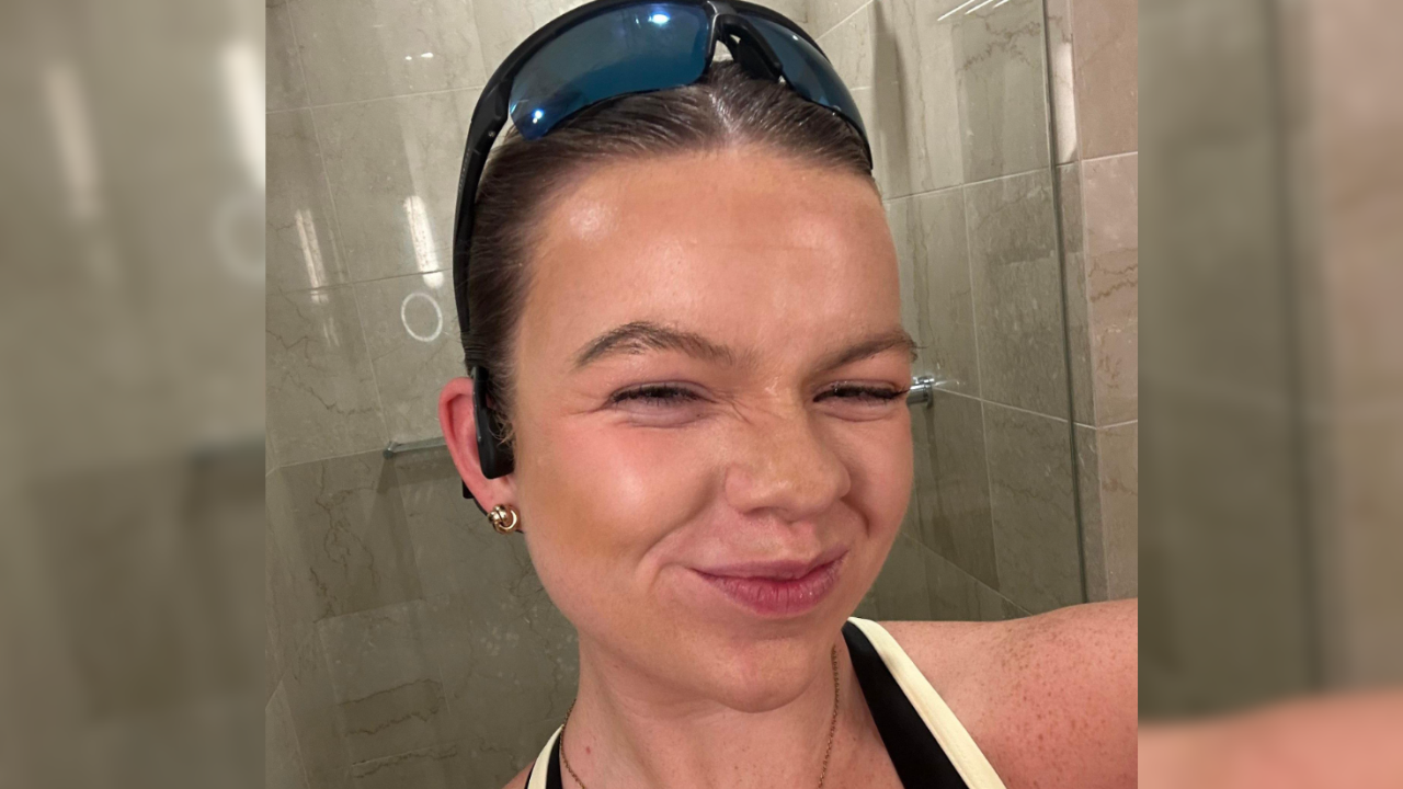 The image is a selfie of a woman smiling, with blue sunglasses on her head and a gold necklace around her neck. A glass shower block can be seen behind her and a small shower head in the top of the photo.